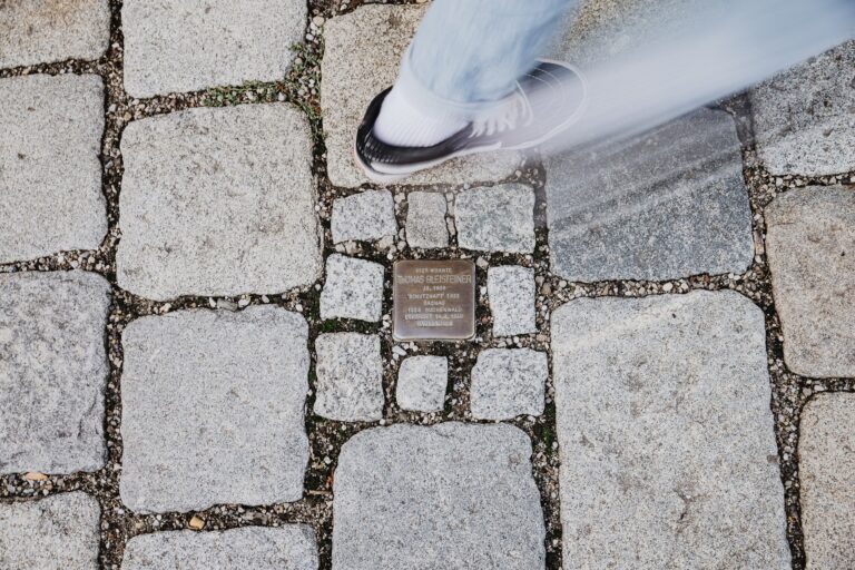 Blick Richtung Boden auf einen Stolperstein eingebettet in Kopfsteinpflaster, ein Fußgänger ist mit eine Fuß in der Bewegung abgebildet. Der Stolperstein gedenkt Thomas Bleisteiner