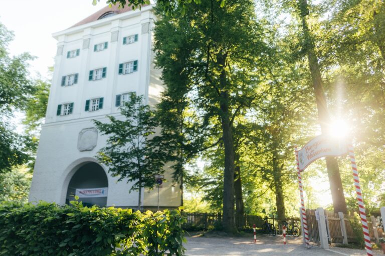Blick auf den Wasserturm, heutige Galerie des Wasserturm e.V. neben den Hofgarten, umgeben von hohen Bäumen, Daneben das Eingangsschild zum Biergarten am Wasserturm. Durch die Bäume blitzten Sonnenstrahlen.