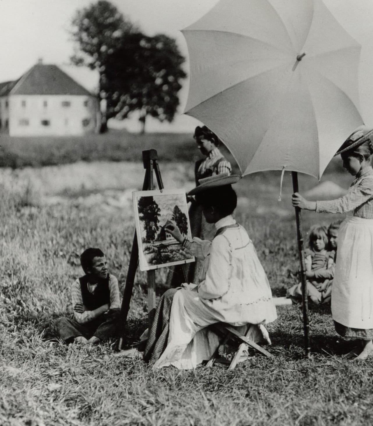 Sogenannte "Malweiber", weibliche Malerinnen um 1900. Die Dame sitzt im hellen Kittel vor der Stafflei und malt. Um sie herum Kinder, ein Mädchen hält ihr einen Sonnenschirm.