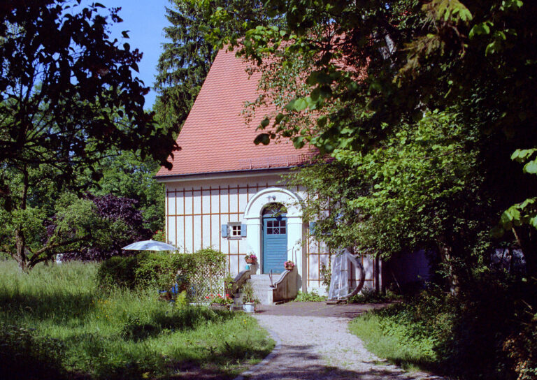 Blick vom Gartenzaun durch den Garten auf das Künstlerhaus Carl Thiemann, welches halb verdeckt hinter Büschen und Bäumen im Sonnenlicht steht. auf der kleinen Treppe zur Eingangstür stehen seitlich Blumentöpfe mit Blühpflanzen. Ein Sonnenschirm ist seitlich aufgespannt.