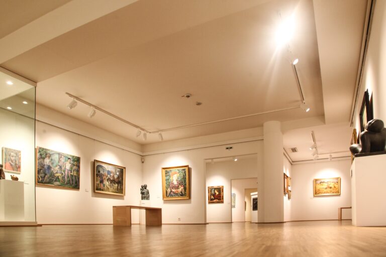 Blick aus Bodennähe in einen Ausstellungsraum der Gemäldegalerie Dachau. An den Wänden sind Gemälde ausgestellt, Skulpturen auf Sockeln und Durchgänge zu weiteren Ausstellungsräumen sind zu sehen.