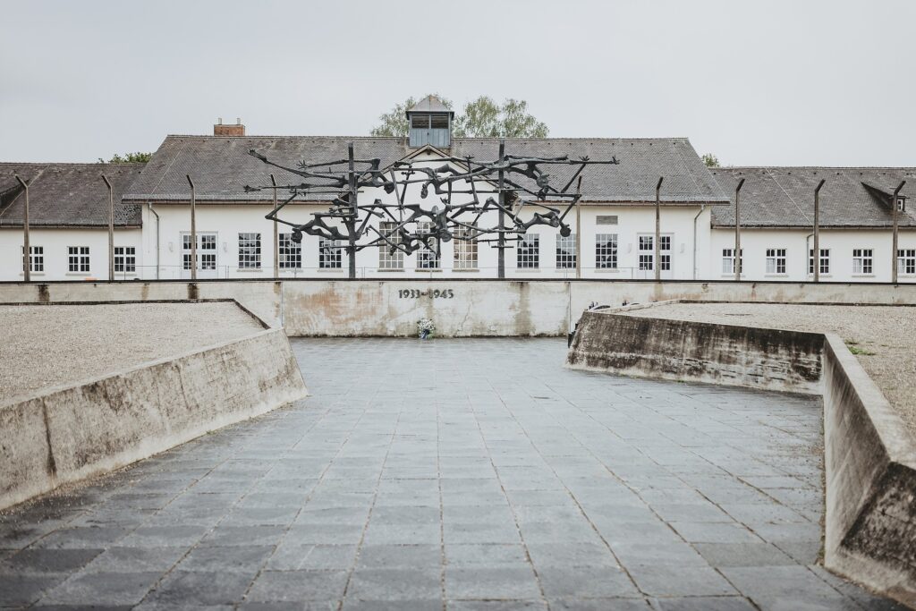 Ansicht des Internationalen Mahnmals auf dem Gelände der KZ-Gedenkstätte Dachau vom ehemaligen Appellplatz aus. Im Hintergrund ist das Jourhaus zu erkennen. Auf einer Mauer unterhalb der Skulptur steht 1933 - 1945, unter den Jahreszahlen ist ein Gedenkkranz aus Blumen mit einer Gedenkschleife an die Wand angelehnt.