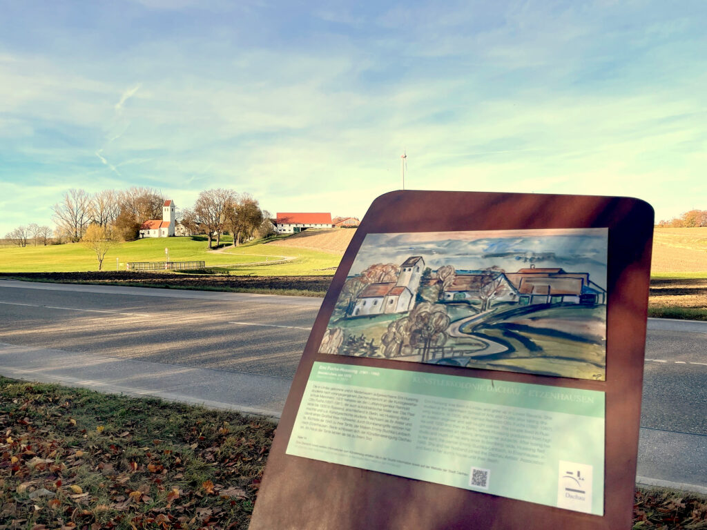 Im Vordergrund angeschnitten eine Metallstele des Künstlerwegs Etzenhausen, dahinter die aktuelle Ortsansicht von Steinkirchen.