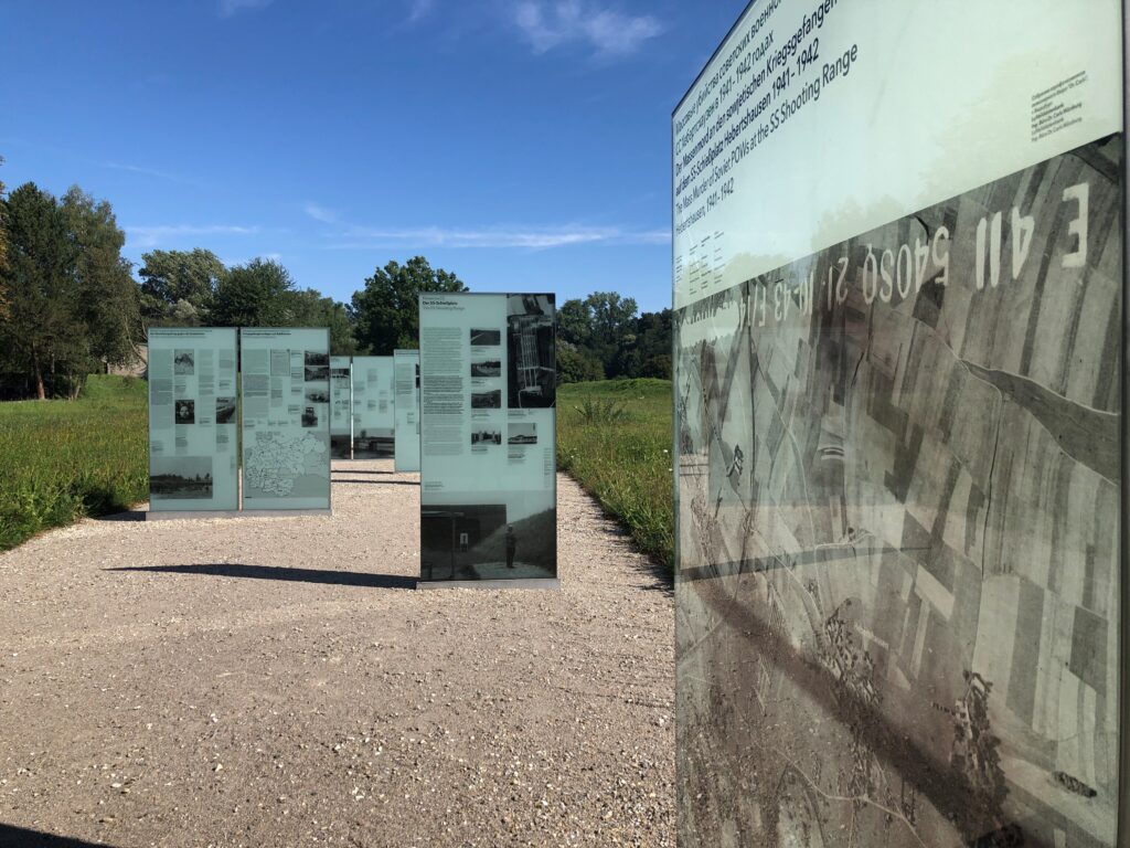 Blick auf mehrere versetzt hintereinander frei stehende mannshohe Gedenktafeln auf einem Kiesplatz an der ehemaligen SS Schießstätte Hebertshausen.
