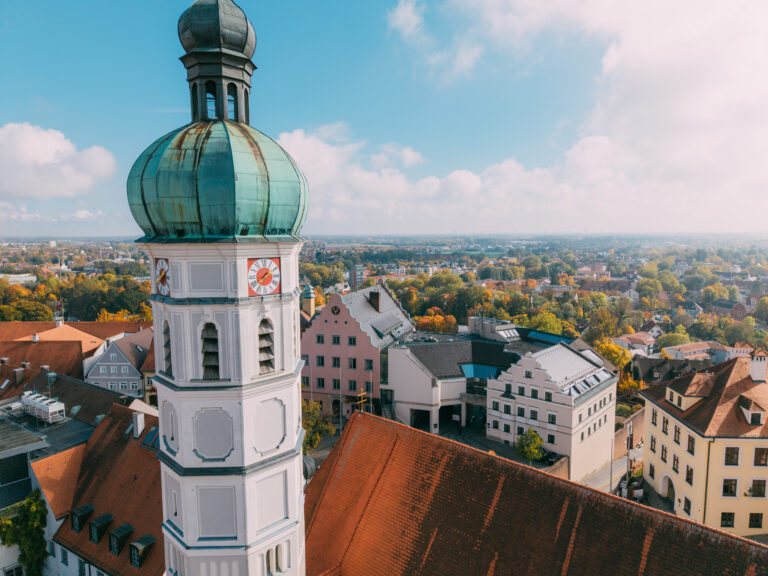 Drohnenaufnahme Blick an der Kirchturmspitze von St. Jakob vorbei auf Konrad-Adenauer-Straße sowie Fassaden und Dächer von altem und neuem Rathaus. Dahinter schweift der Blick in die Ferne über die Dächer Dachaus und die Ebene Richtung München. Milde Herbstsonne und weiße Wolken vor blauem Himmel. Die Bäume zeigen schon Herbstlaub.