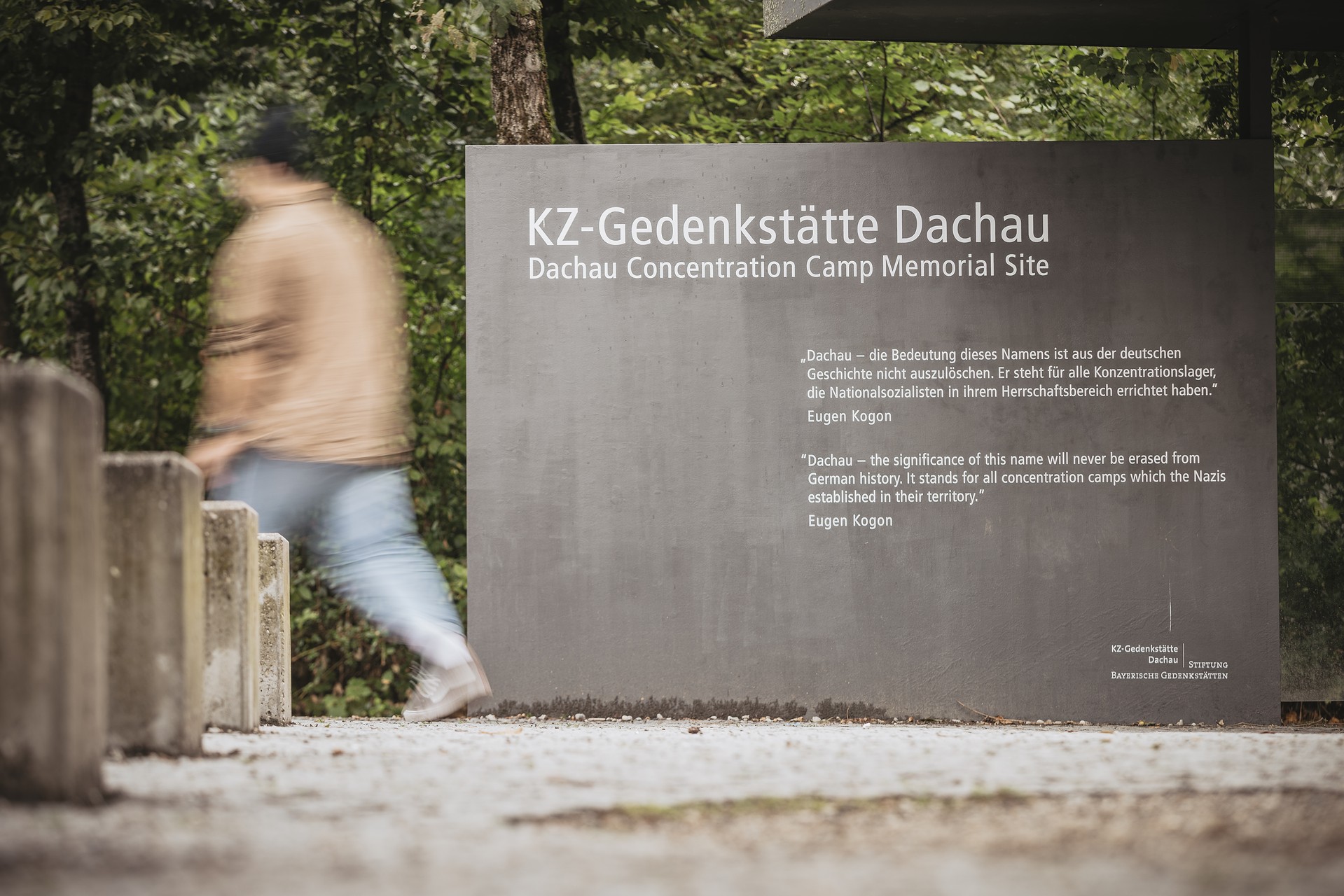 Blick auf Tafel am Eingang der KZ-Gedenkstätte mit der Aufschrift KZ-Gedenkstätte Dachau und Dachau Concentration Camp Memorial Site, darunter noch Zitat Eugen Kogon in Deutsch und Englisch.