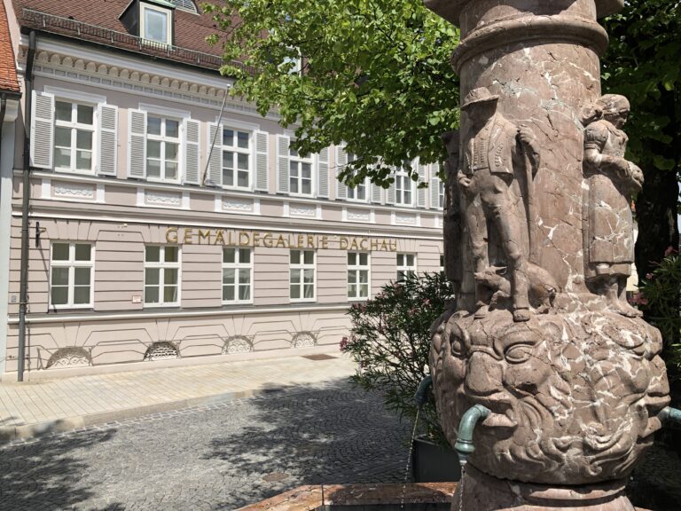 Blick auf einen Ausschnitt der Säule des Ignaz-Taschner-Brunnens auf dem Platz vor dem Rathaus und die Fassade der Gemäldegalerie Dachau im Hintergrund. An der Säule sind über Löwenköpfen zei Figuren abgebildet ein Mann in Dachauer Tracht mit einem Ferkel zwischen den Beinen, eine Frau mit Dachauer Tracht.