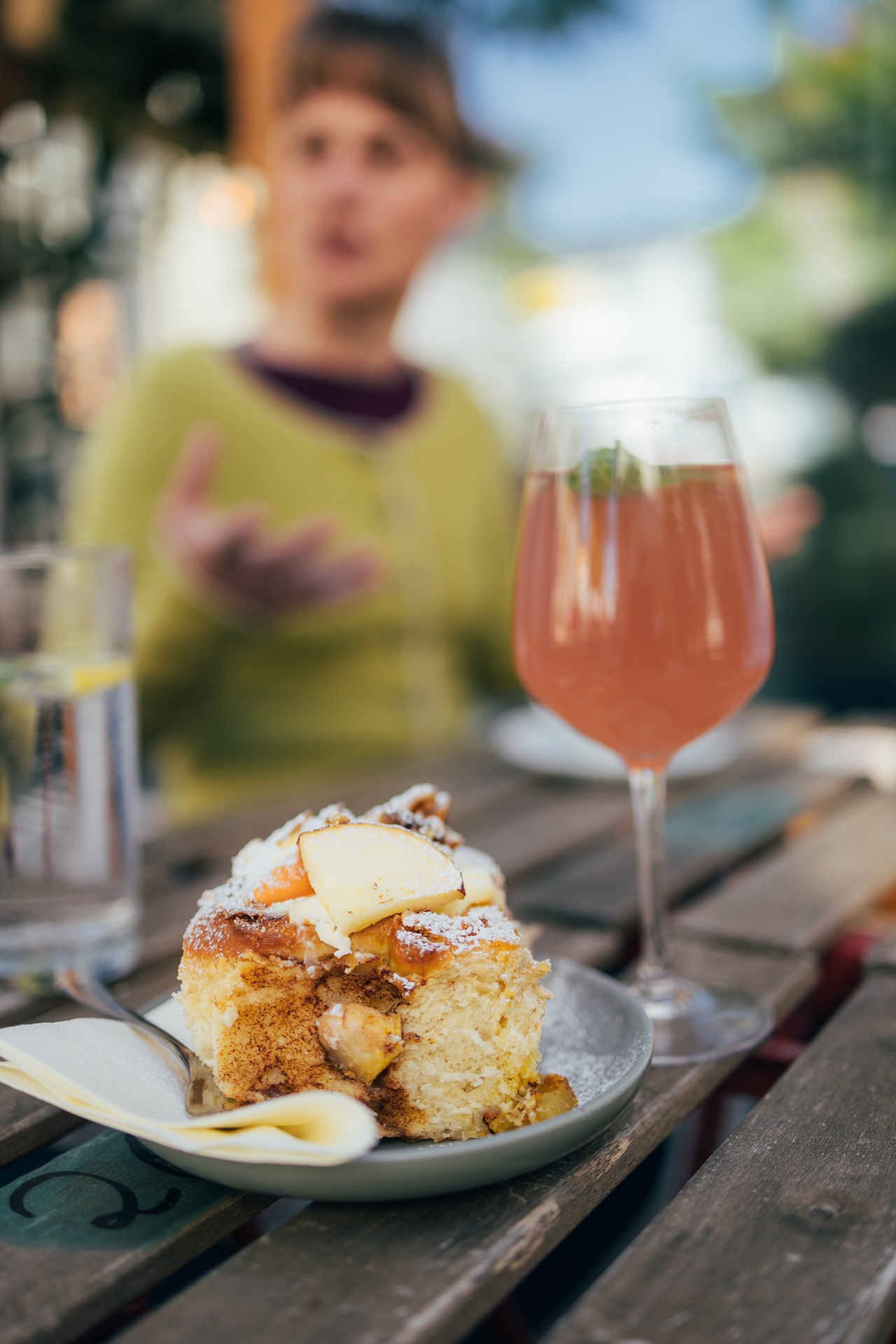 Ein Dachauer Café, auf dem Holztisch steht eine Zimtschnecke und ein Glas mit einem orange rötlichen Sommerdrink. Im Hintergrund ist eine Person zu sehen, die sich unterhält.