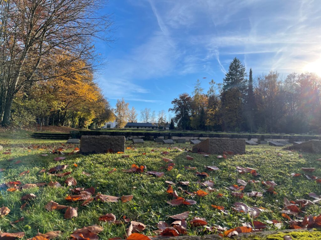 Blick aus Bodenhöhe Richtung terrassenförmig angelegter Gräber und Aussegnungshalle auf dem Walfriedhof Dachau. Auf der Wiese zwischen den Grabsteinen der ehemaligen KZ-Häftlinge und Opfer der Todesmärsche liegt Herbstlaub.