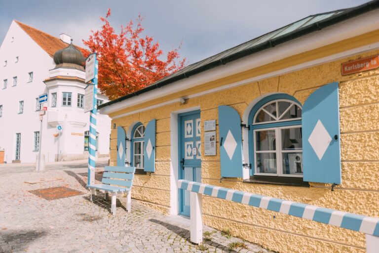 Strahlend sonnige Aufnahme von historischem Zollhäuschen, hellgelbe Fassade und Holzfensterläden in hellblau und weiß sowie einer blauweiß gestreiften Barke (früher zum Anbinden der Pferde), vor dem Häuschen steht eine hellblau/weiße Holzbank, links ist ein blauweiß gestreifter Holzpfosten mit historisch anmutenden Schildern zuerkennen, die auf die ehemalige Zollstation des Marktes Dachau hinweisen.