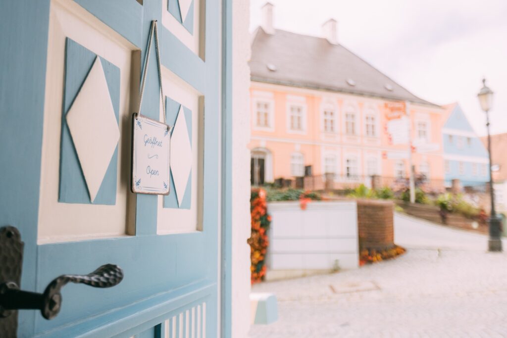 Blick aus der nach innen geöffneten Eingangstür des Zollhäuschens über den kopfsteingepflasterten Karlsberg auf das Bezirksmuseum Dachau und angrenzende bunte Bürgerhäuser auf der Augsburger Straße. An der geöffneten Tür hängt ein farblich passendes und leicht historisch anmutendes Schildchen mit der Aufschrift: "Geöffnet".